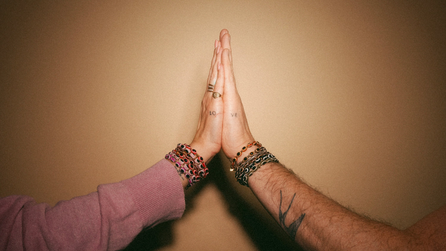 Two hands with tattoos and bracelets held together against a beige background
